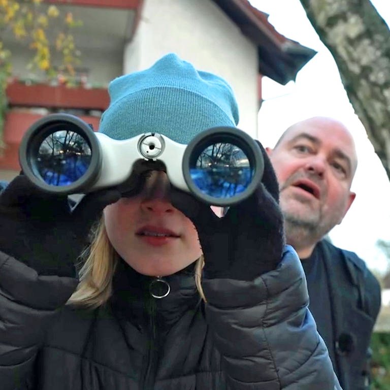 Reporterin Jule schaut durch ein Fernglas - hinter ihr steht Detektiv Alexander